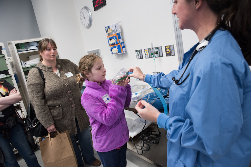 A nurse shows a patient scheduled for spinal fusion surgery an oxygen mask while the patient's mother looks on.