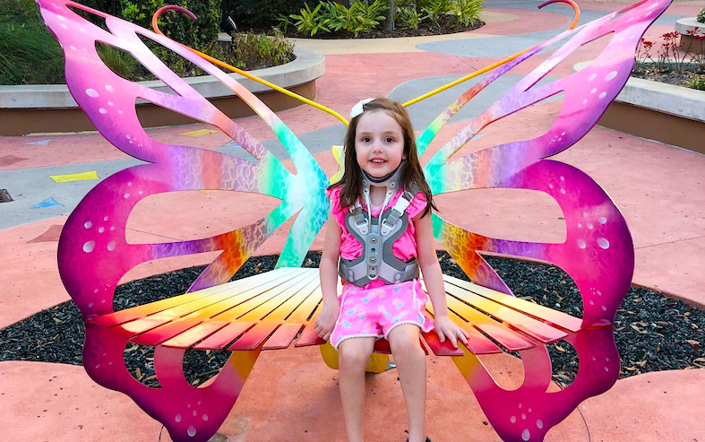 Gracie, wearing a brace after spinal fusion surgery, sits on a bright, pink butterfly bench.