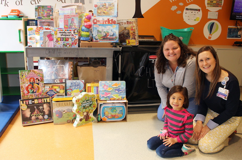 Kalea, who had moyamoya surgery, poses with her clinicians in front of toys she raised for a playroom at the hospital