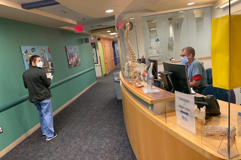 A clinician in the orthopedic urgent care unit sits at a desk behind plexiglass as another walks past. Both are also wearing masks to prevent the spread of COVID-19.
