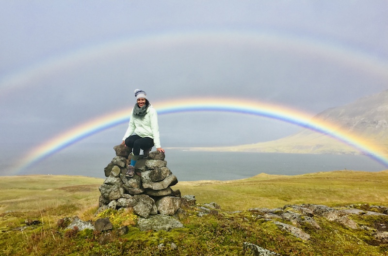 Carla Coutinho in a hat and scarf, poses on a rock structure with a double rainbow in the sky behind her.
