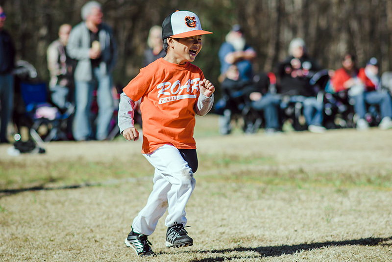 Steven, who has an anorectal malformation runs on a baseball field. He is wearing an orange Little League uniform and laughing.