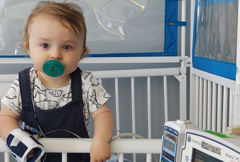 Nathan, who has OMS, stands up in his crib in the hospital. He has a green pacifier in his mouth.